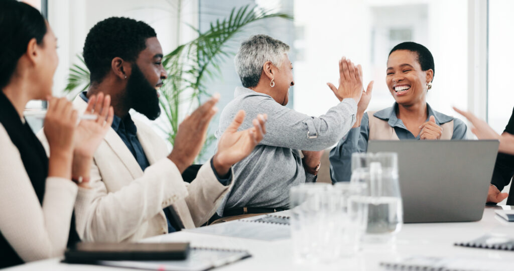 A group of people sitting at a desk in a brightlly lit conference room, cheering the success of the company
